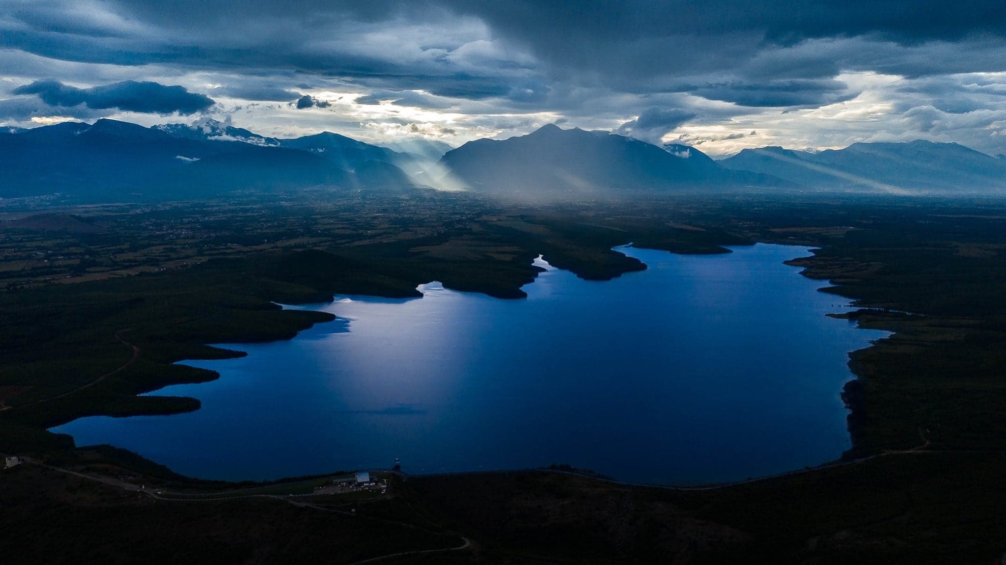 An aerial view of Radoniq Lake under heavy clouds, with rays of light falling on the mountains behind.