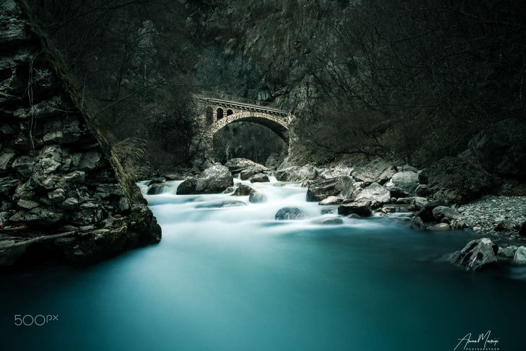 A long exposure of a turquoise river flowing over rocks beneath an old stone arch bridge in a dark gorge.
